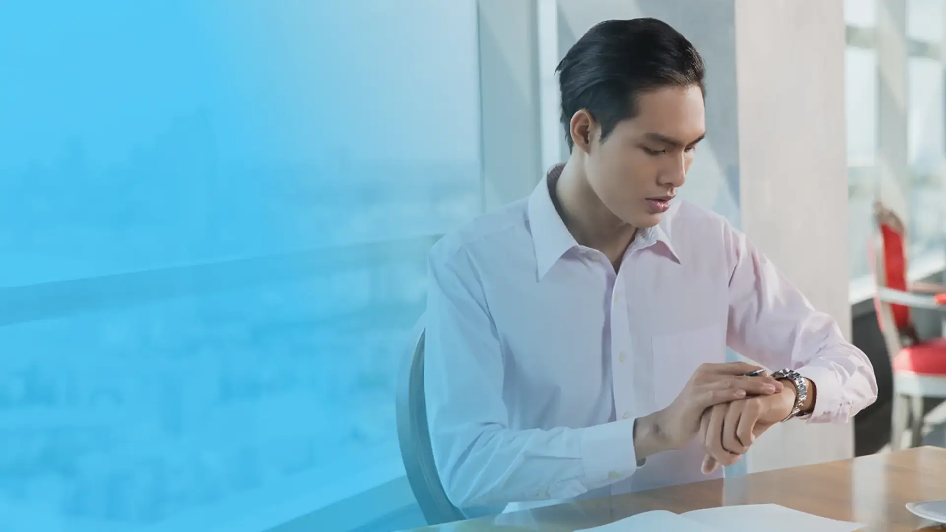 A professional man in a light pink dress shirt sitting at a desk, checking his wristwatch, symbolizing efficient attendance tracking and time management in the workplace.